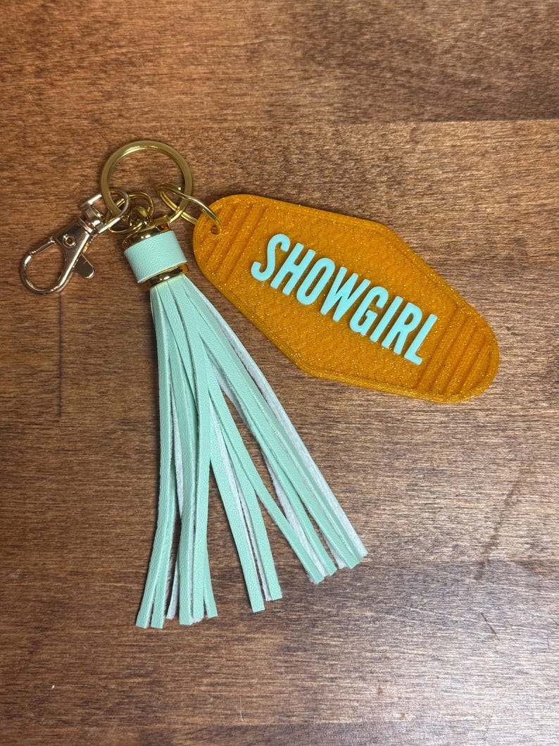 May include: A gold-colored motel-style keychain with the word "SHOWGIRL" in turquoise. The keychain has a gold ring, a mint green tassel, and a lobster clasp.