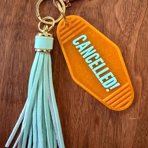 May include: Orange motel-style keychain with the word "CANCELLED!" in light blue. The keychain has a gold-colored clasp and ring, and a light blue tassel.