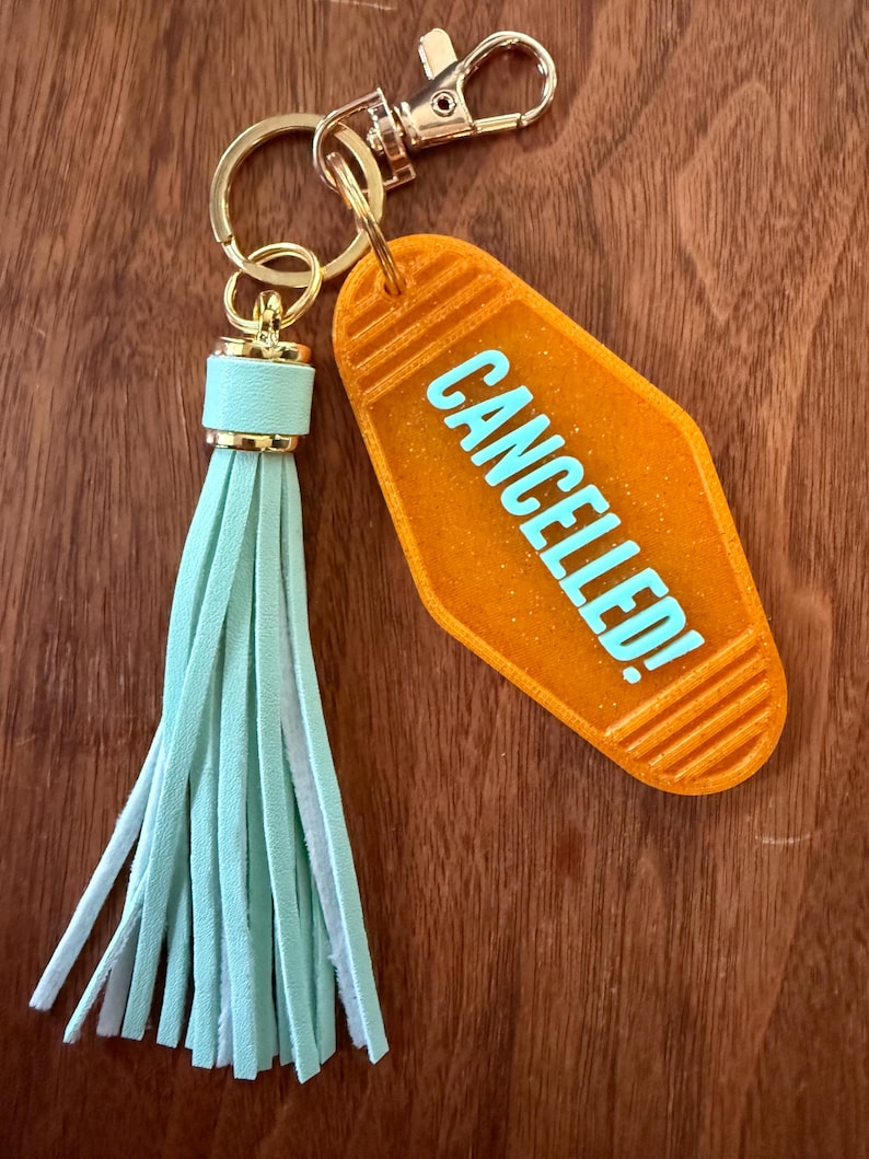May include: Orange motel-style keychain with the word "CANCELLED!" in light blue. The keychain has a gold-colored clasp and ring, and a light blue tassel.