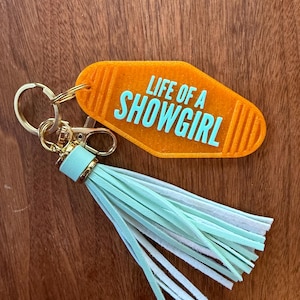 May include: An orange motel-style keychain with the text "LIFE OF A SHOWGIRL" in turquoise. The keychain has a gold-colored clasp and ring, and a tassel with mint green and white fringe.