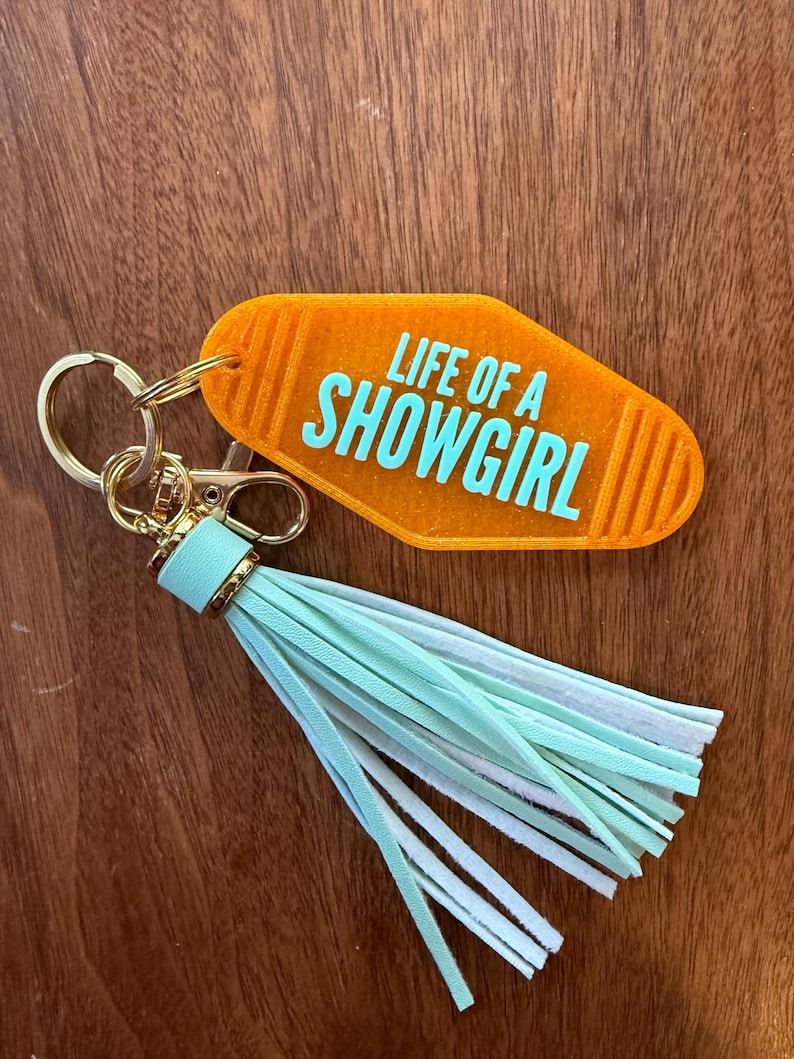 May include: An orange motel-style keychain with the text "LIFE OF A SHOWGIRL" in turquoise. The keychain has a gold-colored clasp and ring, and a tassel with mint green and white fringe.