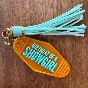 May include: Orange motel-style keychain with the text "BIRTHDAY OF A SHOWGIRL" in turquoise. The keychain has a gold-colored ring and a mint green tassel.