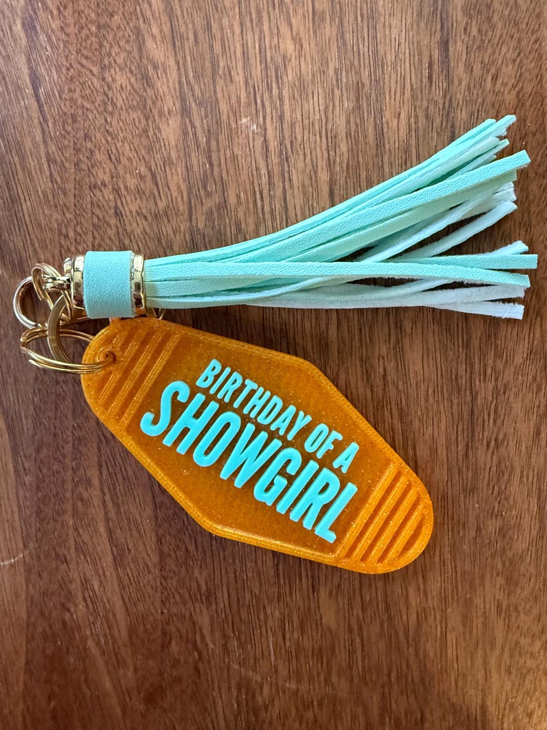 May include: Orange motel-style keychain with the text "BIRTHDAY OF A SHOWGIRL" in turquoise. The keychain has a gold-colored ring and a mint green tassel.