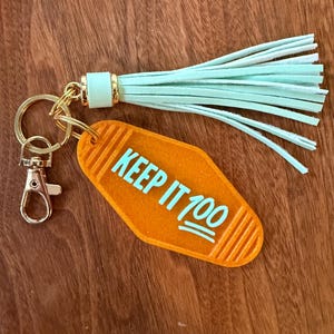 May include: Orange motel-style keychain with the phrase "KEEP IT 100" in turquoise. The keychain has a gold-colored clasp and ring, and a mint green tassel. The background is a wooden surface.