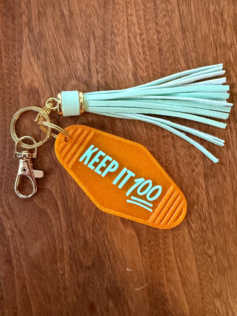 May include: Orange motel-style keychain with the phrase "KEEP IT 100" in turquoise. The keychain has a gold-colored clasp and ring, and a mint green tassel. The background is a wooden surface.