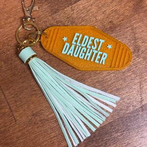 May include: An orange motel-style keychain with the words "ELDEST DAUGHTER" in turquoise. The keychain has a gold-colored clasp and ring, and a mint green and white tassel.