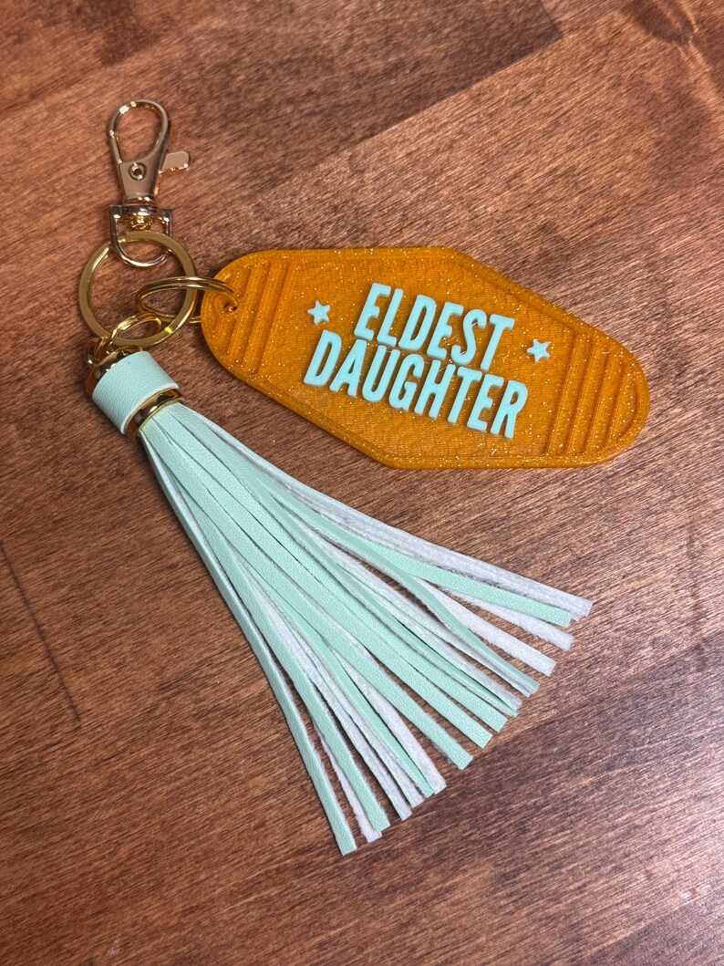 May include: An orange motel-style keychain with the words "ELDEST DAUGHTER" in turquoise. The keychain has a gold-colored clasp and ring, and a mint green and white tassel.