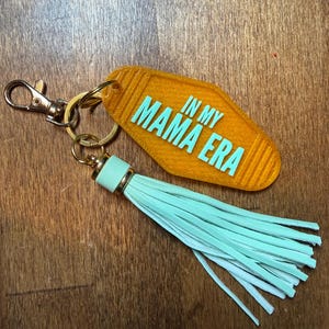 May include: An orange motel-style keychain with the text "IN MY MAMA ERA" in turquoise. It features a gold-colored clasp and ring, and a light turquoise tassel. The keychain is likely made of acrylic or a similar material.