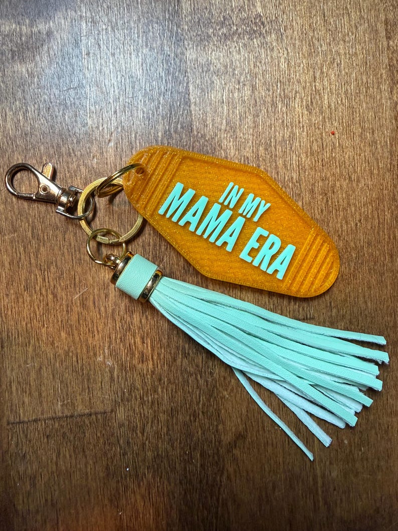 May include: An orange motel-style keychain with the text "IN MY MAMA ERA" in turquoise. It features a gold-colored clasp and ring, and a light turquoise tassel. The keychain is likely made of acrylic or a similar material.