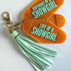 May include: Orange motel-style keychains with the phrases "BIRTHDAY OF A SHOWGIRL" and "LIFE OF A SHOWGIRL" in mint green. The keychains are attached to a gold-colored clasp and a mint green tassel.