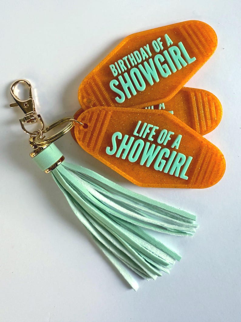 May include: Orange motel-style keychains with the phrases "BIRTHDAY OF A SHOWGIRL" and "LIFE OF A SHOWGIRL" in mint green. The keychains are attached to a gold-colored clasp and a mint green tassel.