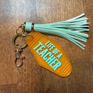 May include: An orange keychain with the text "LIFE OF A TEACHER" in turquoise. The keychain has a gold ring, a clasp, and a mint green tassel.