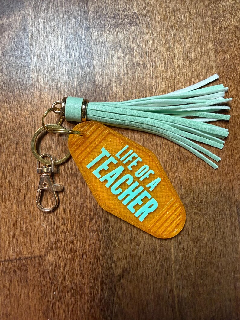 May include: An orange keychain with the text "LIFE OF A TEACHER" in turquoise. The keychain has a gold ring, a clasp, and a mint green tassel.