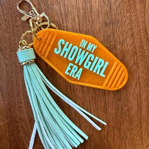 May include: An orange motel-style keychain with the text "IN MY SHOWGIRL ERA" in turquoise. It features a gold-colored clasp and ring, and a mint green tassel. The keychain is set against a wooden background.