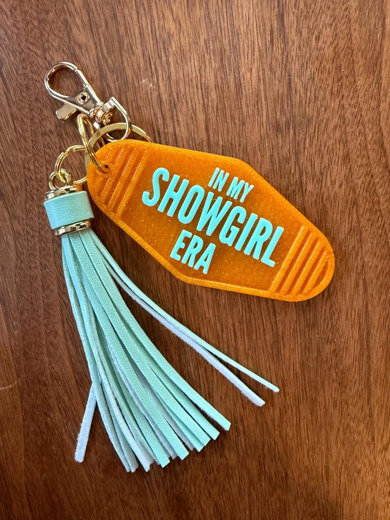 May include: An orange motel-style keychain with the text "IN MY SHOWGIRL ERA" in turquoise. It features a gold-colored clasp and ring, and a mint green tassel. The keychain is set against a wooden background.
