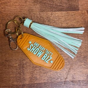 May include: A keychain with a retro motel-style tag in orange with the text "AND BABY THAT'S SHOW BIZ" in turquoise. It has a gold-colored keyring and clasp, and a mint green tassel.