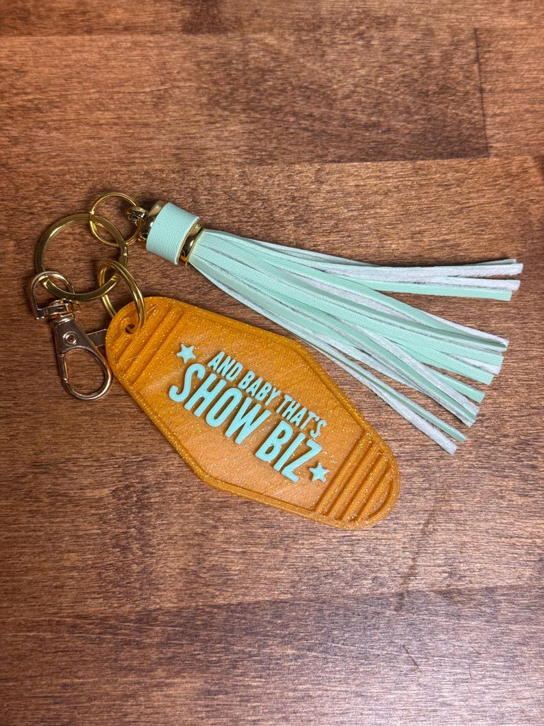 May include: A keychain with a retro motel-style tag in orange with the text "AND BABY THAT'S SHOW BIZ" in turquoise. It has a gold-colored keyring and clasp, and a mint green tassel.