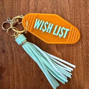 May include: An orange motel-style keychain with the words "WISH LIST" in turquoise. The keychain has a gold-colored clasp and a mint green tassel. The background is a wooden surface.