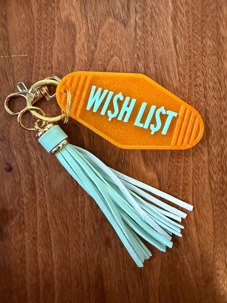 May include: An orange motel-style keychain with the words "WISH LIST" in turquoise. The keychain has a gold-colored clasp and a mint green tassel. The background is a wooden surface.