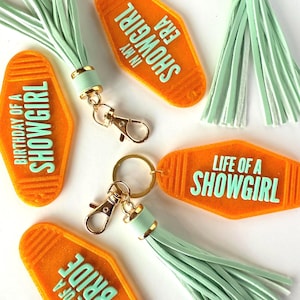 May include: Orange motel-style keychains with mint green tassels and gold hardware. The keychains have text that reads "Life of a Bride", "Birthday of a Showgirl", "In My Showgirl Era", and "Life of a Showgirl".