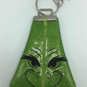 May include: Green glittery keychain pouch featuring a cartoon character face with black embroidered details and a zipper closure. The keychain has a silver metal ring and clasp.