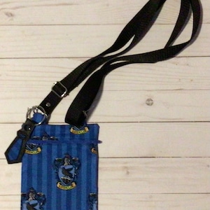 Puede incluir: Una funda para teléfono de tela azul y amarilla con una correa negra. La funda presenta un patrón repetido del escudo de la casa de Hogwarts para Ravenclaw, que incluye un cuervo y las palabras "Ravenclaw".