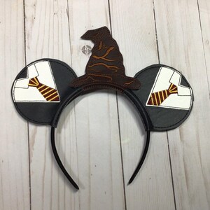 May include: Black mouse ears headband with a brown Sorting Hat and two black ears. Each ear features a white shirt and a gold and burgundy striped tie. A fun accessory for fans of the wizarding world.