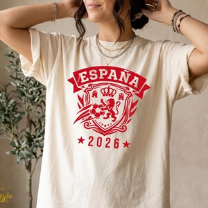 May include: A cream-colored short-sleeve t-shirt featuring a red graphic print on the chest. The design includes the text "ESPAÑA" above a shield emblem containing a lion, a crown, and a soccer ball, with the year "2026" flanked by two stars below.