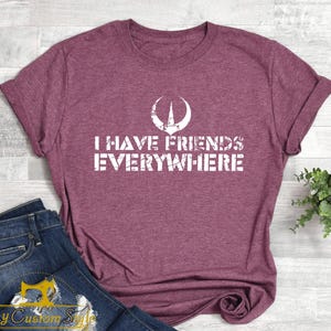 May include: Maroon t-shirt with rolled sleeves and the text "I HAVE FRIENDS EVERYWHERE" in white block letters. A white graphic is above the text. The shirt is paired with blue jeans.