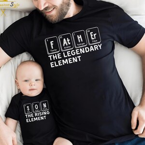 May include: Black t-shirts with white text. One shirt reads "FATHER THE LEGENDARY ELEMENT" with periodic table elements. The other shirt reads "SON THE RISING ELEMENT". The shirts are worn by a father and baby.