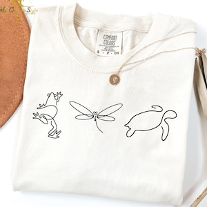 May include: Cream-colored t-shirt featuring minimalist line art of a frog, dragonfly, and turtle. The shirt has a crew neck and short sleeves. A gold necklace with a pendant is draped across the shirt. The tag reads "Comfort Colors".