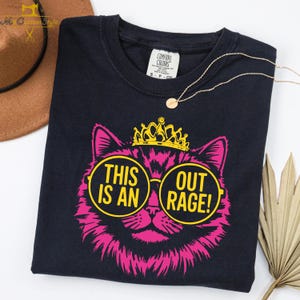 May include: A navy blue t-shirt featuring a pink cat wearing yellow-rimmed glasses and a crown. The text "THIS IS AN OUTRAGE!" is displayed in yellow circles. A gold necklace and a Comfort Colors tag are also visible.