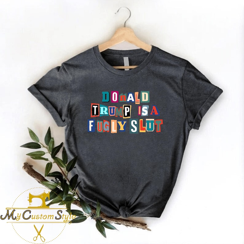 May include: Dark gray t-shirt with the text "DONALD TRUMP IS A FUGLY SLUT" in colorful block letters. The shirt is hanging on a wooden hanger against a white background. A sprig of greenery is placed on the lower left.