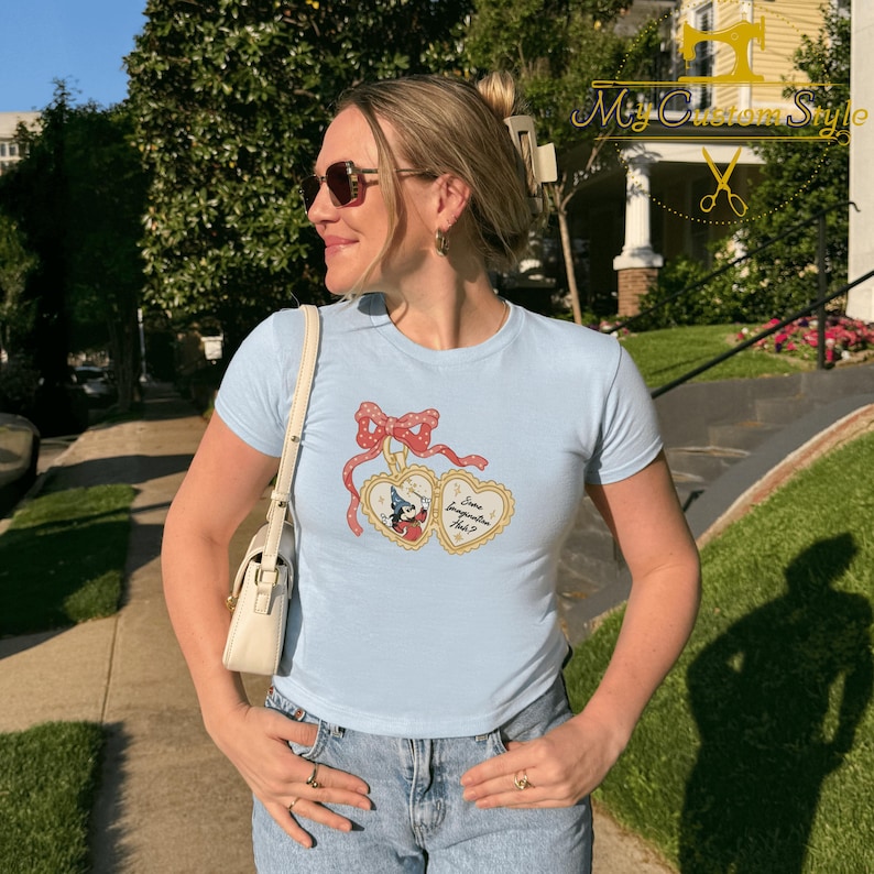 May include: A light blue t-shirt featuring a graphic of two heart-shaped lockets with a red bow and ribbon. The lockets have a cartoon character inside. The text on the shirt reads "Some Imagination Ash?". The person is wearing sunglasses and jeans.