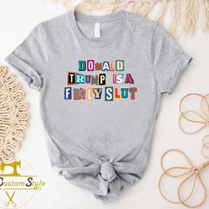 May include: A light gray t-shirt with the text "DONALD TRUMP IS A FUGLY SLUT" in colorful block letters. The shirt is knotted at the waist. The logo "My Custom Style" is visible in the lower left corner.