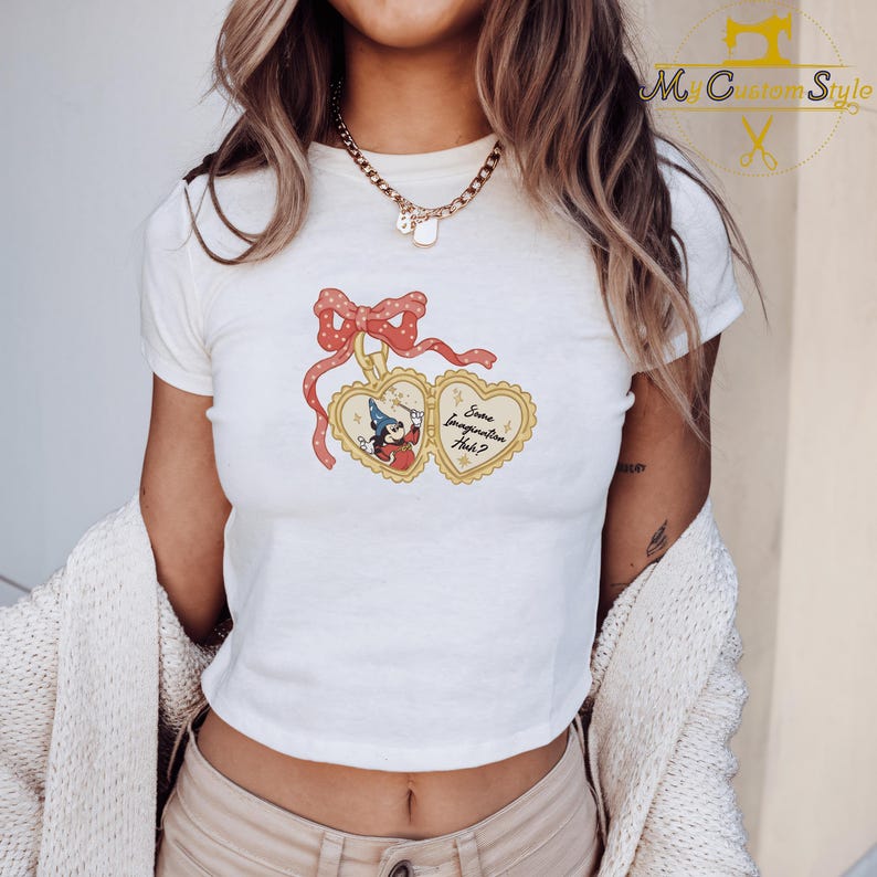 May include: White crop top featuring a whimsical graphic of a heart-shaped locket with a cartoon character and the text "Some Imagination Huh?" The locket is adorned with a red bow and ribbons. The shirt has short sleeves.