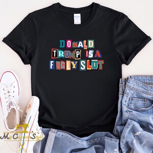 May include: Black t-shirt with the text "DONALD TRUMP IS A FUGLY SLUT" in colorful, block-letter style. The shirt is paired with blue jeans and white sneakers. The shirt is a Gildan brand.