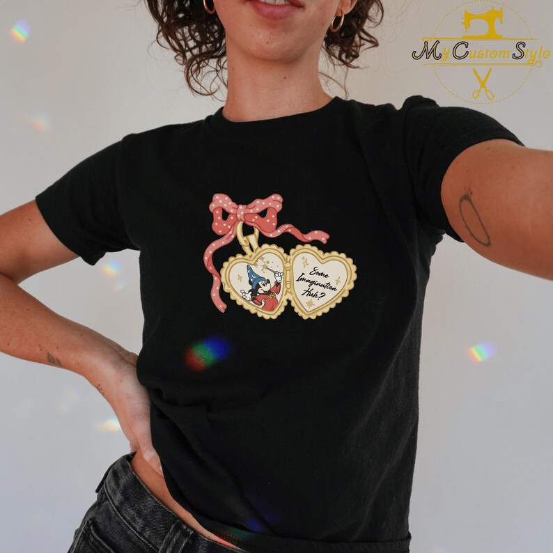 May include: Black t-shirt featuring a heart-shaped pendant design with a pink bow and the text "Some Imagination Huh?". The pendant has a cartoon character with a magic wand. The shirt is a crew neck.