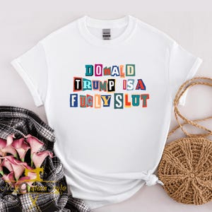 May include: White t-shirt with the text "DONALD TRUMP IS A FUGLY SLUT" in colorful block letters. The shirt is made of a soft material and has short sleeves. The text is in a variety of colors, including blue, red, and yellow.