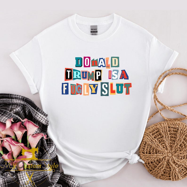 May include: White t-shirt with the text "DONALD TRUMP IS A FUGLY SLUT" in colorful block letters. The shirt is made of a soft material and has short sleeves. The text is in a variety of colors, including blue, red, and yellow.