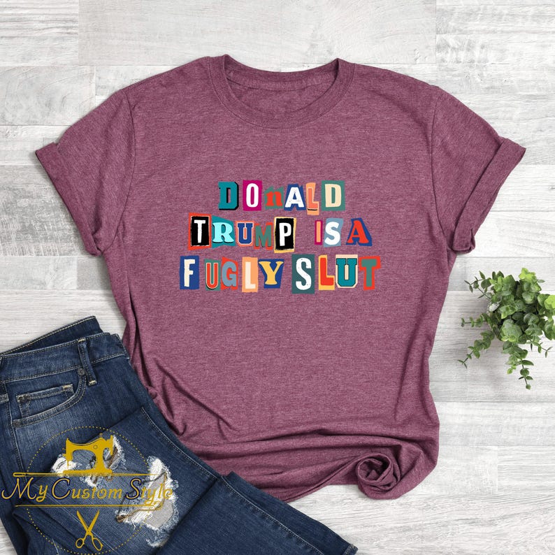 May include: Maroon t-shirt with the text "DONALD TRUMP IS A FUGLY SLUT" in colorful block letters. The shirt is paired with blue jeans. A small plant is visible in the background.
