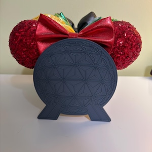 May include: A pair of mouse ears with red sequined ears, a red satin bow, and a dark blue geometric design. The bow has a rainbow-colored accent. The ears are designed to be worn on the head.