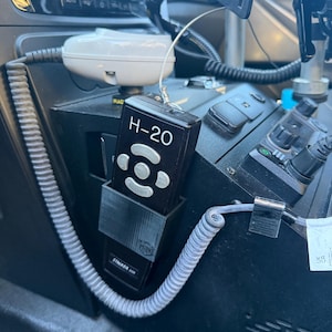 May include: A black H-20 Stalker radar remote control unit housed in a custom-fitted black plastic mount. The remote features a central directional pad with four buttons and a coiled grey cable extending from the base, secured within a vehicle console.