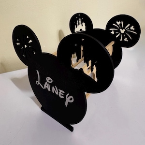 Può includere: Scaffale nero in legno a forma di Mickey Mouse con il nome "Laney" in corsivo bianco. Lo scaffale presenta disegni ritagliati di un castello e fuochi d'artificio nelle orecchie. Lo scaffale è in legno e ha un interno in legno chiaro.