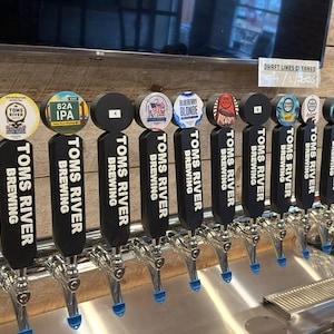 May include: A row of beer taps with various tap handles. The handles are black with the words "Toms River Brewing" and different beer names. The tap handles have colorful circular labels with beer names and logos.