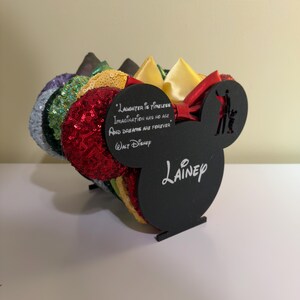 May include: A display of colorful sequined Mickey Mouse ear headbands. The black display stand features the quote "Laughter is Timeless Imagination has no age And dreams are forever" and the name "Lainey".