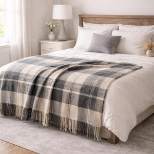 Può includere: Una coperta a quadri grigia e crema con frange, drappeggiata su un letto con lenzuola bianche e un cuscino grigio. La camera da letto ha una palette di colori neutri con una struttura letto in legno e un comodino bianco.