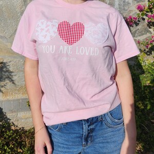 May include: A pale pink t-shirt featuring three heart motifs. The central heart is red gingham, with white hearts on either side, patterned with bows and lace. The text reads "YOU ARE LOVED" and "1 JOHN 4:19".