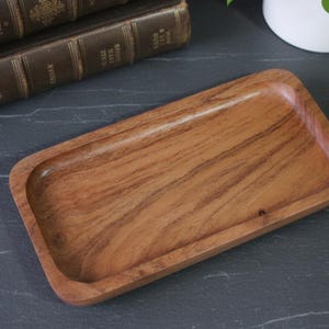 May include: A rectangular wooden tray with rounded corners. The tray is made of light brown wood with visible grain patterns. The tray is empty and sits on a dark grey surface.