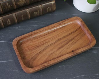 May include: A rectangular wooden tray with rounded corners. The tray is made of a light brown wood with visible grain patterns. The tray is empty and sits on a dark gray surface.